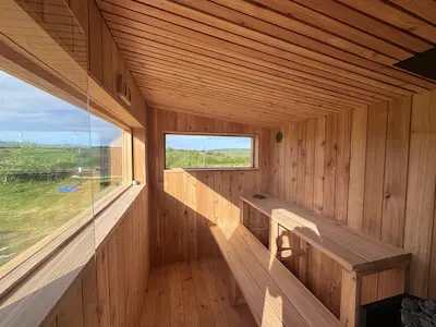 The interior of a modern cedar wood sauna features tiered benches and large windows overlooking a green landscape
